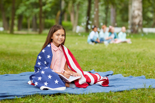 Full length portrait of cute girl covered by American flag sitting on picnic blanket in park and smiling at camera, copy space