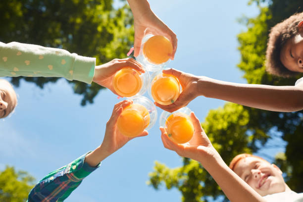 Kids Cheering with Orange Juice Low angle close up of kids holding glasses with orange juice against blue sky outdoors, copy space juice drink stock pictures, royalty-free photos & images