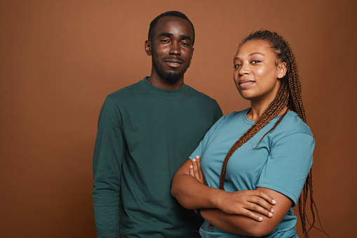 Minimal waist up portrait of modern African-American couple looking at camera while standing confidently against brown background in studio, copy space