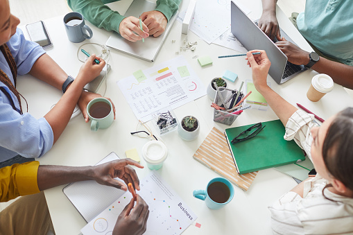 High angle view at contemporary creative team working together at cluttered table with coffee mugs and documents, teamworking or studying concept, copy space