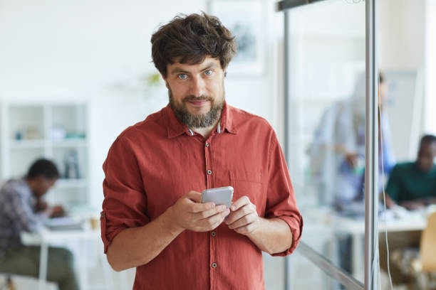istock Smiling Bearded Man Holding Smartphone in Office 1266963388