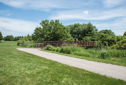 Path to footbridge surrounded by native grasses and deciduous trees under a blue sky with clouds at the natural reserve in Aurora, Illinois