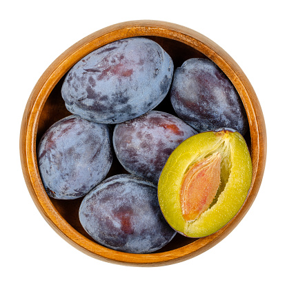Ripe European plums with cross section of one fruit in a wooden bowl. Freestone fruit with purple, violet and black skin, also called Zwetschge or Damson. Prunus domestica. Close up from above. Photo.