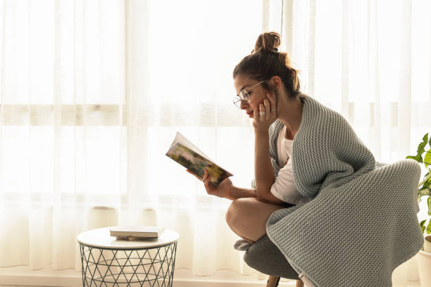 young beautiful girl with reading glasses sitting in the chair near window reading the book with natural sunlight and enjoy, with film grain - woman with glasses reading a book imagens e fotografias de stock