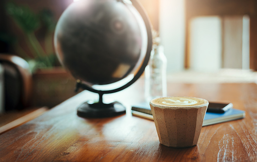 Close-up latte coffee in ceramic cup on wooden table with blurred  simulated globe and notebook on background, Vintage light and tone