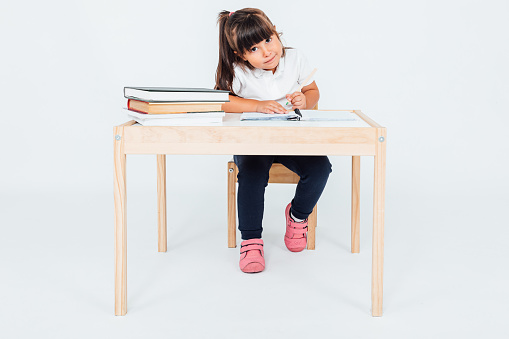 Brunette girl at school, smiling sitting on a chair next to a table with books painting, on white background. School concept