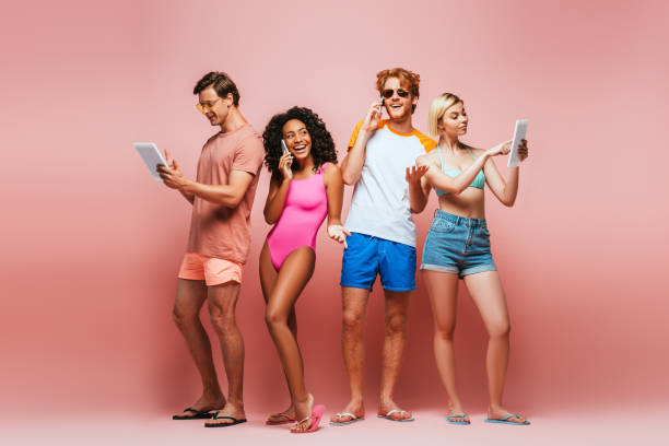 полная длина зрения молодых мультикультурных друзей в летний наряд с использованием цифровых устройств на розовый - shorts women people young women стоковые фото и изображения