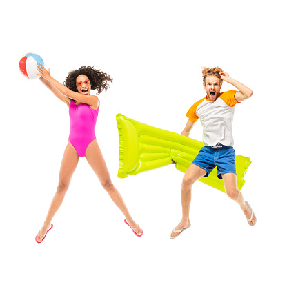 Excited multiethnic friends holding inflatable ball and mattress while jumping isolated on white