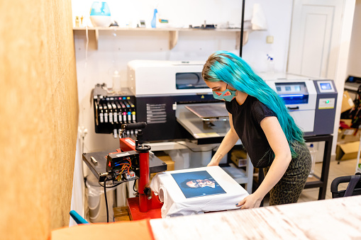 Young female business owner , with dyed blue hair printing t-shirt in the silk screen printing machine at her workshop while wearing protective face mask during COVID-19