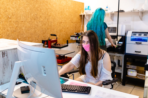 Woman with Friedreich ataxia sitting in wheelchair and receiving online orders, while her colleague working on a digital prinitng machine in their workshop while both wearing protective face mask during COVID-19 pandemic