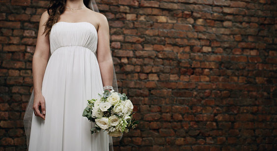 Cropped shot of an unrecognisable woman holding a bouquet against a brick wall on her wedding day