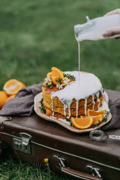 Homemade carrot cake stands on a wooden plate on a vintage suitcase. The cake is topped with a sweet white sauce. The dessert is decorated with orange slices and fresh flowers.Orange dessert.