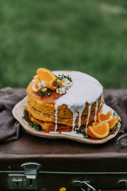 Homemade carrot cake stands on a wooden plate on a vintage suitcase. The cake is covered with white sweet cream. The dessert is decorated with orange slices and fresh flowers.Orange dessert.