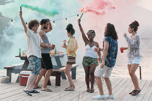 Group of happy friends dancing on a pier together among colored smoke