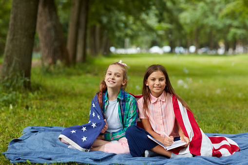 Full length portrait of two cute girls covered by American flag sitting on picnic blanket in park and smiling at camera, copy space