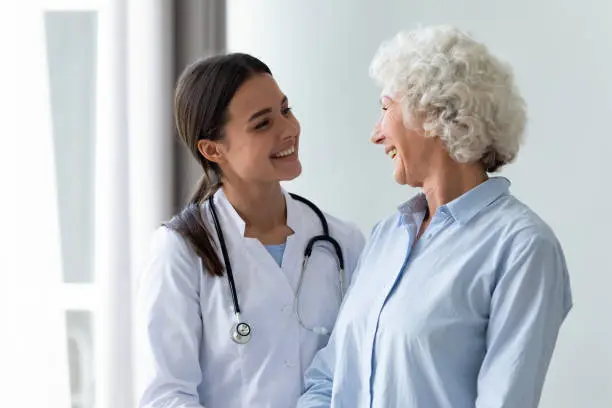 Smiling young female general practitioner enjoying sincere talk, sharing good health news with hoary older senior woman indoors. Caring millennial nurse helping middle aged patient at meeting.