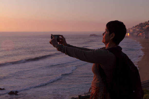 Woman at the beach photographing the sunset Latin athletic senior active woman at the beach photographing a beautiful sunset with her cellphone using 5G Technology wireless at Reñaca, Viña del Mar, Valparaiso, Chile. vina del mar chile stock pictures, royalty-free photos & images