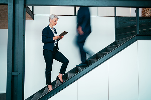 Shot of an attractive businesswoman using digital tablet while walking up stairs in busy office. Two business people in a motion on stairs in the office.