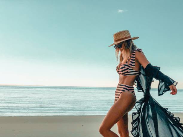 Beautiful woman at the beach in a stripped pinky bikini wearing a cover-up with mesh transparencies swimwear dress and a straw hat by the sea photography - fotografia de stock