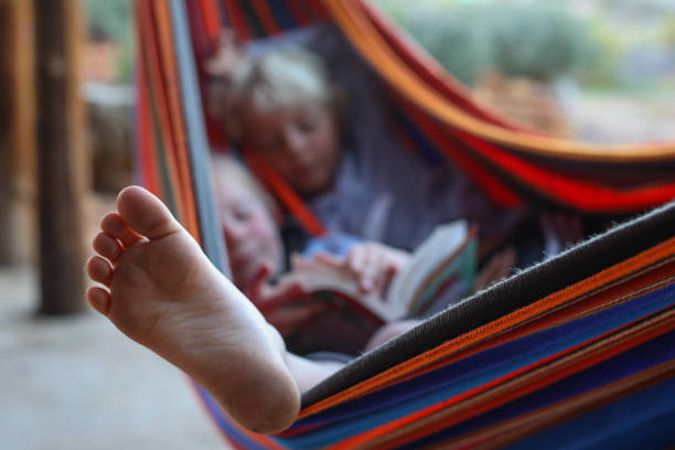 Two brothers are lying together in a hammock while reading from the same paperback book. The focus is on the one boys foot hanging out of the hammock.