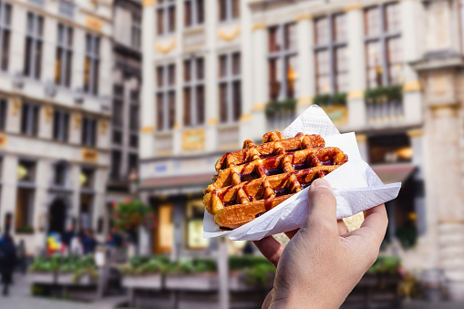 Man holds in hand popular street food - Traditional Belgian dessert - tasty waffle with chocolate sauce on the background of city tourist streets of  Brussels, Belgium, Europe. Traditional Belgian dessert.