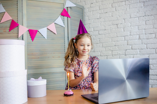 Children celebrating her birthday through video call virtual party with friends. Lits out candle. Coronavirus outbreak 2020. Birthday party with cake online together with her friend.