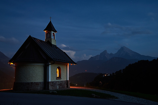 Traditional chapel Kirchleitn illuminated and Mount Watzmann on a beautiful summer evening with blue sky. Alpine landscape and nature during blue hour in national park Berchtesgaden, Bavaria, Germany. August 2020