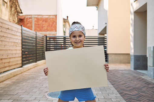 Young cheerful smiling girl holding an empty cardboard sign outdoors in the city in front of a residential building.