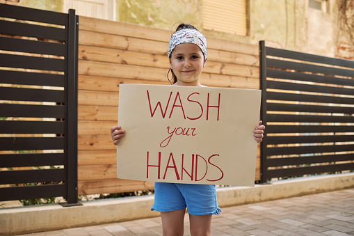 Young cheerful smiling girl holding a wash your hands cardboard sign outdoors in the city in front of a residential building during a coronavirus, covid-19 pandemic.