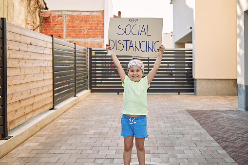 Young cheerful smiling girl holding a keep social distancing cardboard sign outdoors in the city in front of a residential building during a coronavirus, covid-19 pandemic.