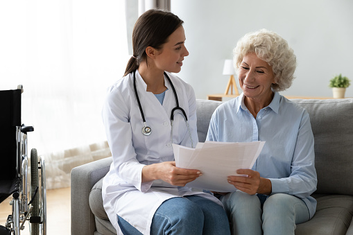 Happy young skilled female general practitioner in white medic uniform visiting disabled smiling middle aged senior patient, showing health analysis on paper, satisfied with treatment results.