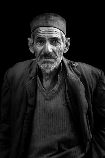 Black and white street portrait of an old man with wrinkles and white beard, standing against black background, staring towards the camera  in the month of June 2019 in Manali, Himachal Pradesh, India