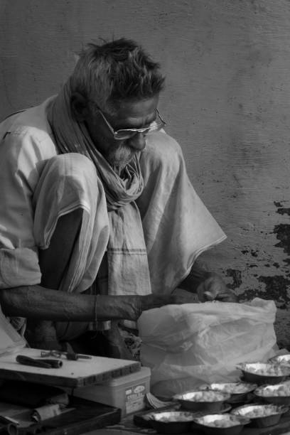 Black and white street photograph of an old man with white beard and glasses, cooking food in the streets while sitting down in the month of December 2018 in Rishikesh, Uttarakhand, India