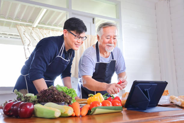 alter asiatischer mann im rentenalter, der zeit damit verbringt, zu lernen, wie man gesundes essen aus dem internet mit seinem sohn mit einer vielzahl von bio-gemüse kocht - senior adult digital tablet domestic life learning stock-fotos und bilder