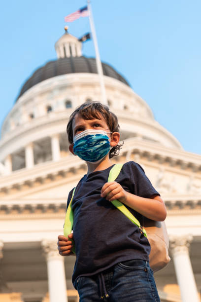 serious little schoolboy wearing a protective face mask and a backpack in front of the capitol building - little boys preschooler back to school backpack imagens e fotografias de stock