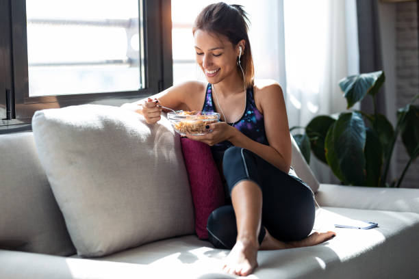 sporty young woman eating a bowl of muesli while listening music sitting on the couch at home. - eating women breakfast cereal imagens e fotografias de stock