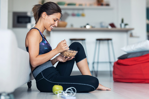 Shot of sporty young woman eating a bowl of muesli while listening music sitting on the floor at home.