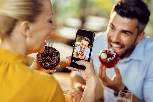 Close-up of couple having fun while eating donuts in a cafe and taking picture with mobile phone. Focus is on man on phone's screen.