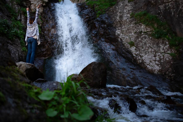 Young happy woman enjoing the waterfall. Woman standing in front of waterfall with rased hands and smile. Concept of ecotourism travel. Waterfall Shumka, Dombai, Karachay-Cherkessia, Russia