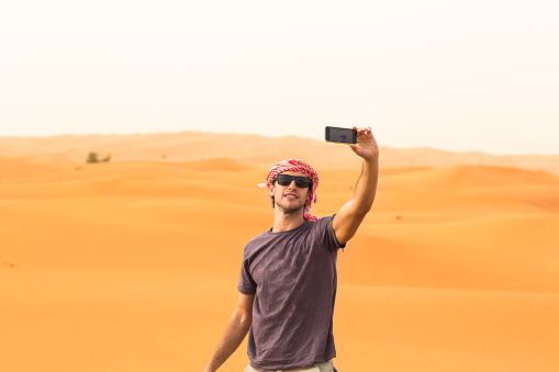 Dubai tourism and travel adventure.Young man using his smartphone to take a picture on top the golden sand dunes in the desert. He is enjoying a safari tour while wearing casual clothes and a local Arab headwear.