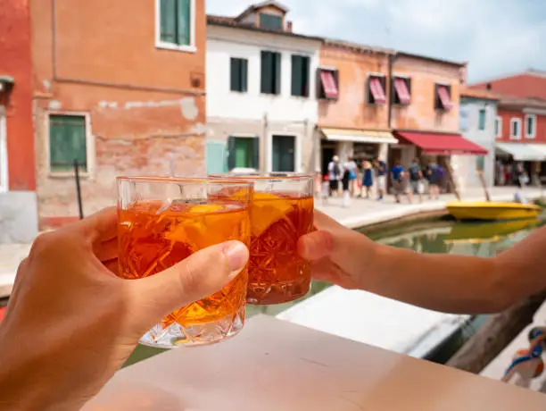 Two Hands hold glasses of spritz cocktails with ice to make cheers in Burano, the city with colorful houses in Venice, Italy