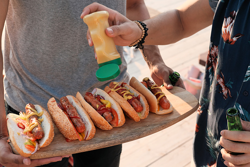 Close-up of young people adding sauce on hot dogs they preparing food for the party outdoors