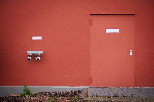 Red concrete wall with red steel door and two fire hydrant pipes, part of an industrial building.