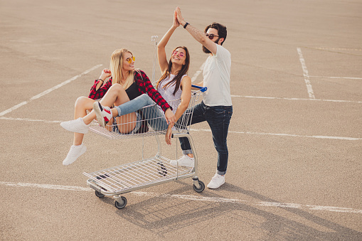 High angle of carefree male and female friends in casual clothes riding shopping cart and giving high five on summer day on parking lot