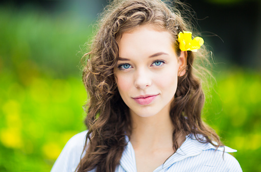 Friendly beautiful young female teenager person with amazing blue eyes and a pretty face. She has a yellow flower on her ear at the park. She is staring at the camera against a green background.