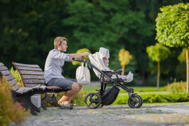 młody człowiek siedzący na drewnianej ławce w miejskim zielonym parku w ciepły, słoneczny letni dzień. dziecko w białym wózku obok ojca. relaks po długim spacerze. widok z boku. spokojna atmosfera w przyrodzie. - caucasian pensive male young adult zdjęcia i obrazy z banku zdjęć