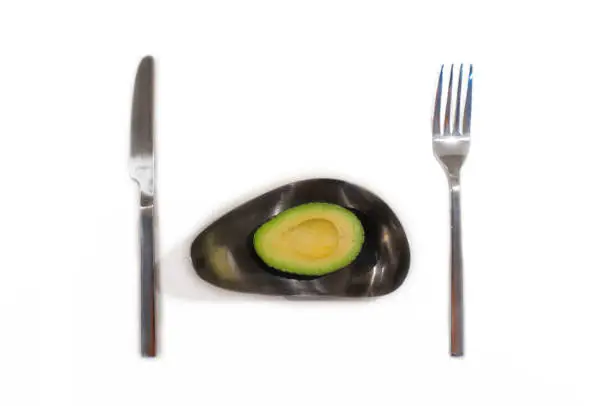 Half an avocado on silver plate with knife and fork isolated on white background.