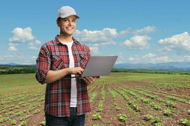 istock Young man in a checkered shirt holding a laptop computer and looking at the camera on a field with plants 1267129927