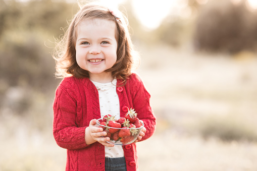 Smiling baby girl 2-3 year old eating fresh ripe strawberry outdoors closeup. Happiness. Healthy lifestyle.