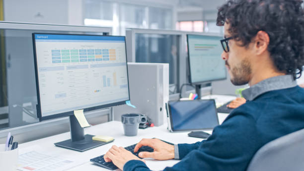 istock Young Handsome Manager with Curly Hair Works on a Desktop Computer and Compares Data with a Software. Diverse and Motivated Business People Work on Computers in Modern Open Office. 1267134380